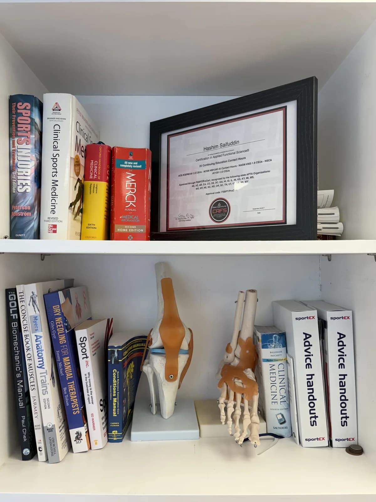 Reference shelf at Atlas Osteopathy Finchley with anatomy models, clinical sports-medicine texts and Hashim's qualifications.