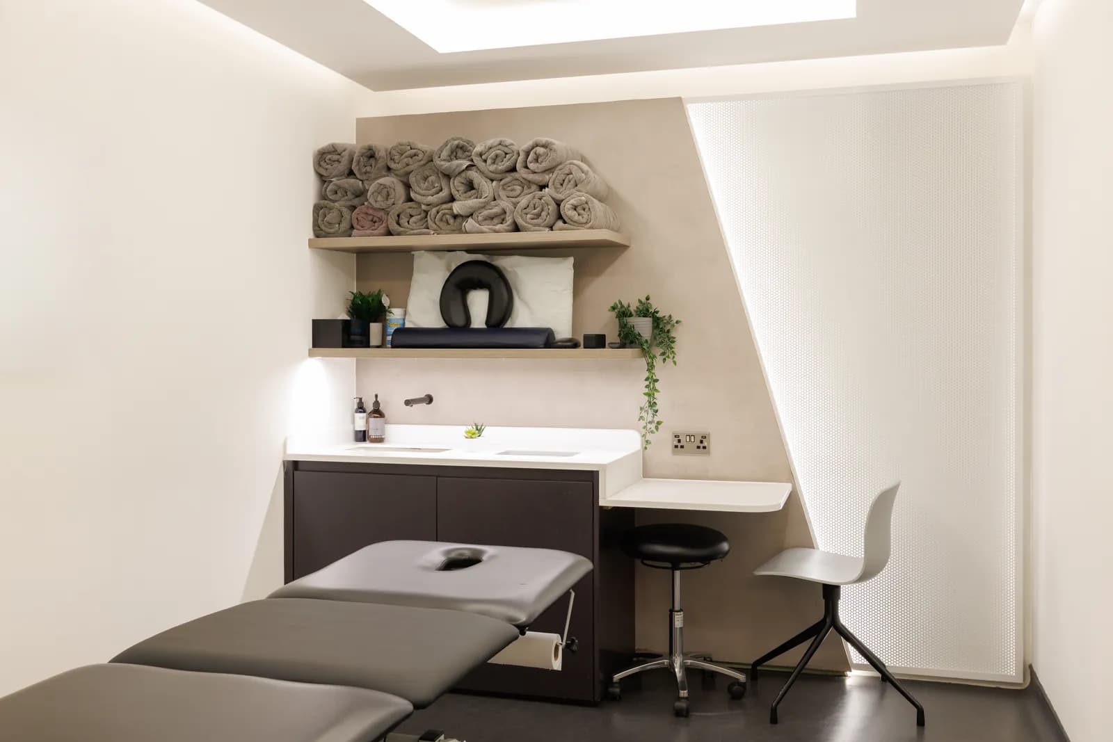 Treatment room at Atlas Osteopathy Liverpool Street with adjustable plinth, rolled-towel shelving, sink and stool.